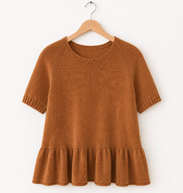 Kate’s Shirt in warmem Hellbraun als Produktfoto vor einer weißen Wand. Das nahtlos von oben gestrickte Shirt zeigt eine schlichte Raglankonstruktion, kurze Ärmel mit 1-rechts-1-links-Bündchen und ein sanft ausgestelltes Rüschenbündchen am Saum für eine feminine Silhouette.