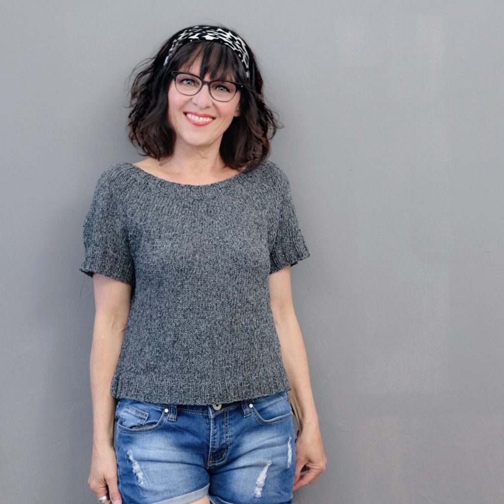 Frau mit Brille trägt das graue Mapala Shirt mit kurzen Ärmeln und Rundpasse zu Jeans, Frontansicht vor grauem Hintergrund.
