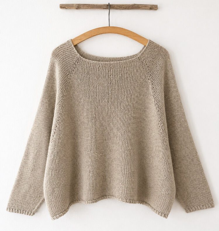 Raakel Tee als handgestricktes Oversize-Shirt mit weiten Fledermausärmeln und Rundhalsausschnitt. Das nahtlos von oben gestrickte Modell hängt auf einem Holzbügel vor einer weißen Wand und zeigt den ruhigen Fall sowie die schlichte, moderne Silhouette.