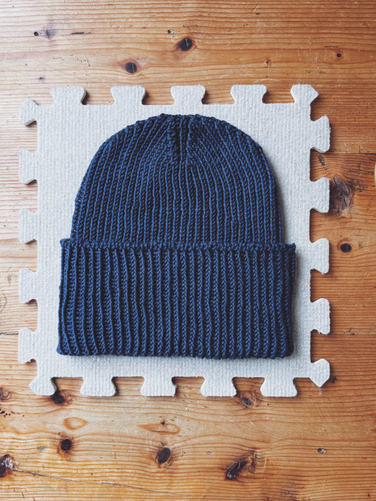 Zoora Hat in Dunkelblau beim Spannen auf der Matte – warme Unisex-Mütze mit Strukturmuster und Umschlag, gestrickt in Runden.