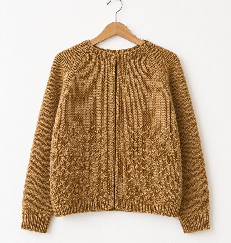Gelber handgestrickter Cardigan mit Raglanärmeln auf einem Holzbügel vor weißer Wand, mit Strukturmuster im unteren Bereich und schlichten Rippenbündchen.