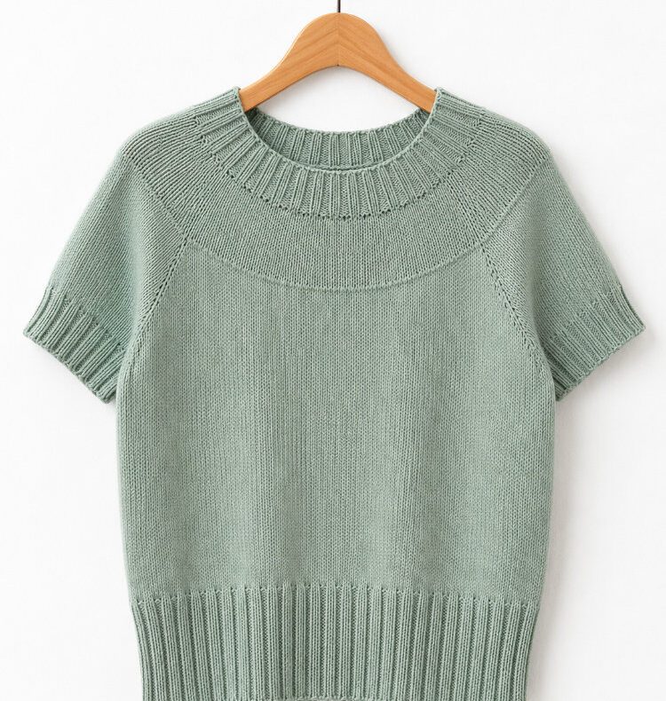 Makikki Shirt aus salbeigrünem Garn als minimalistisches Produktfoto vor weißem Hintergrund. Das nahtlos von oben nach unten gestrickte Shirt zeigt eine runde Passe mit Raglanzunahmen, einen Rundhalsausschnitt, kurze Ärmel mit breiten Bündchen und einen geraden, lässigen Schnitt.