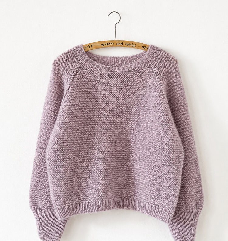 Produktfoto der Piilo Blouse in zartem Rosé an einem Holzkleiderbügel vor einer schlichten weißen Wand. Die nahtlos von oben gestrickte Blouse zeigt ihre markante Raglankonstruktion, den runden Halsausschnitt sowie breite Rippenbündchen an Ärmeln und Saum. Das weiche Garn mit leichtem Halo verleiht dem Modell eine luftige, moderne Optik.