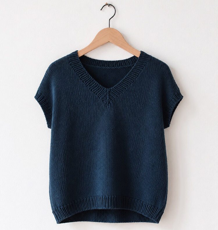 Dunkelblaues handgestricktes Vaatha Tee mit kleinem V-Ausschnitt und angeschnittenen Ärmeln auf einem Holzbügel vor einer weißen Wand. Die schlichte Silhouette und die klare Strickstruktur kommen im weichen Tageslicht besonders gut zur Geltung.