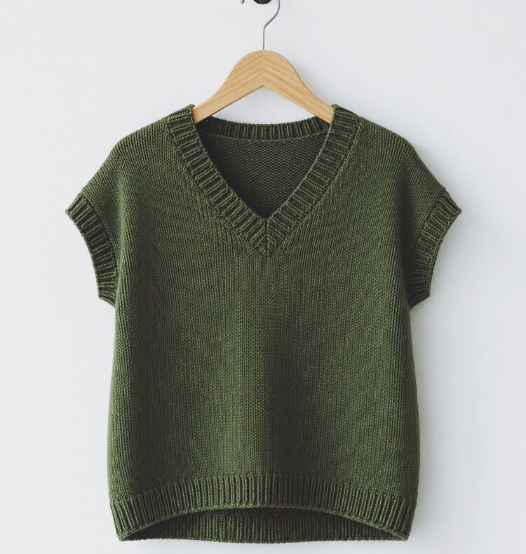 Frau trägt das grüne Vaatha Tee als Pullunder über einem schmalen Langarmshirt und hält eine Tasse in der Hand.