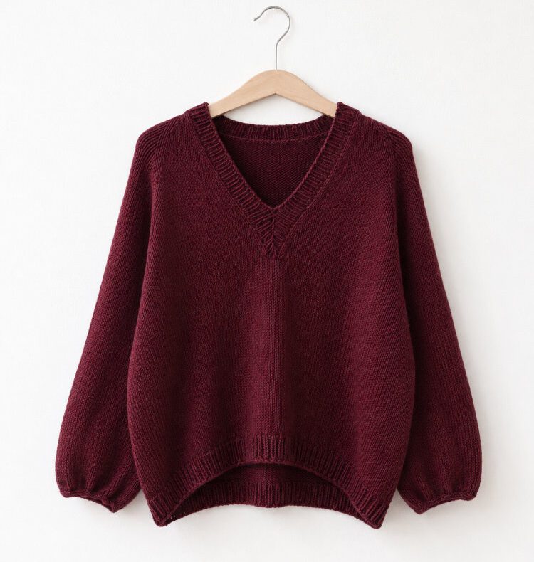 Koora Blouse mit V-Ausschnitt und langen Ärmeln in dunklem Pflaumenrot, gestrickt aus DROPS Saga Mix 21, auf einem Holzbügel vor weißer Wand.