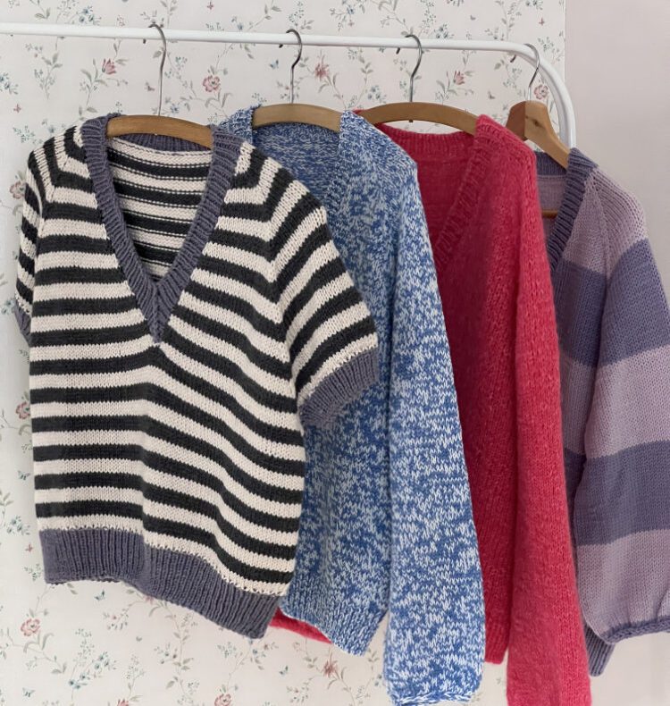 Koora Blouse – gestreifte Bluse mit V-Ausschnitt auf Kleiderbügel, daneben gefaltete Strickteile in Rot und Lila.