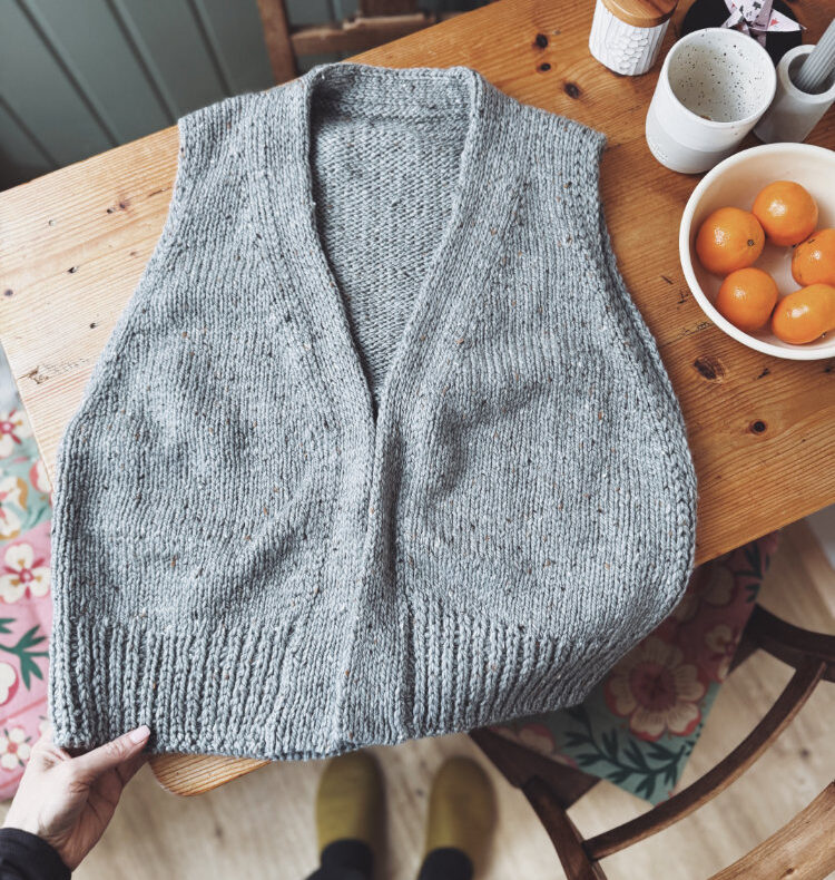 Erikka Vest aus Hamilton Tweed – ärmellose Strickweste mit V-Ausschnitt, nahtlos von oben nach unten gestrickt, fotografiert im Flatlay auf Holztisch mit Garnknäuel und Kaffee.