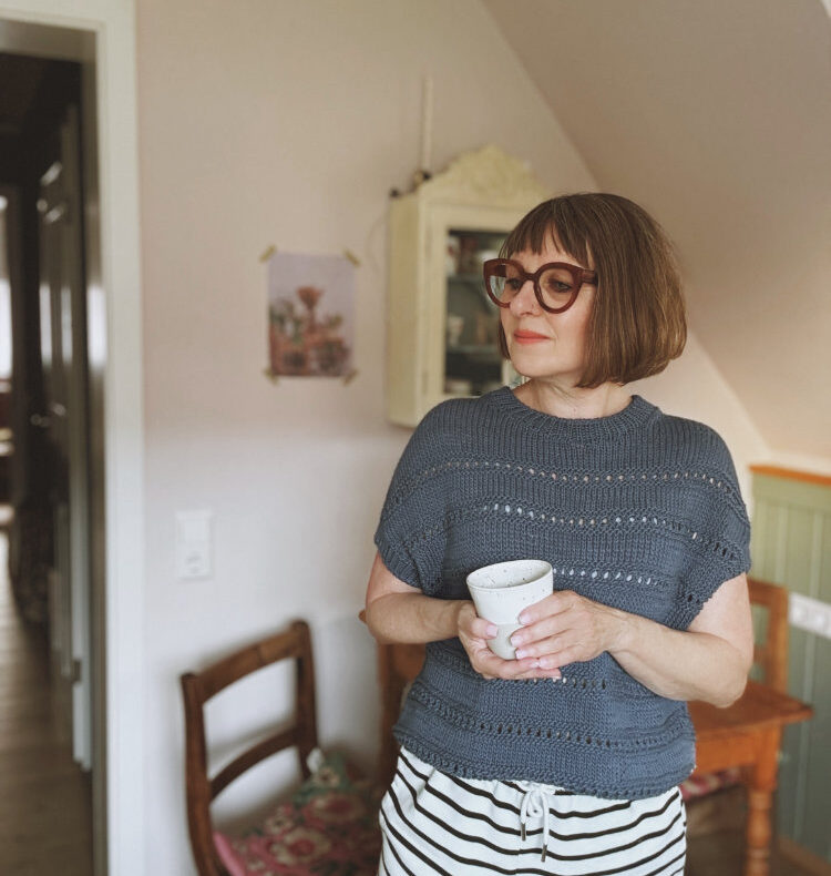 Frau steht in Hytta Tee vor heller Wand – gestricktes Shirt in Pastellblau, glatt rechts, nahtlos von oben gestrickt, Rundhalsausschnitt