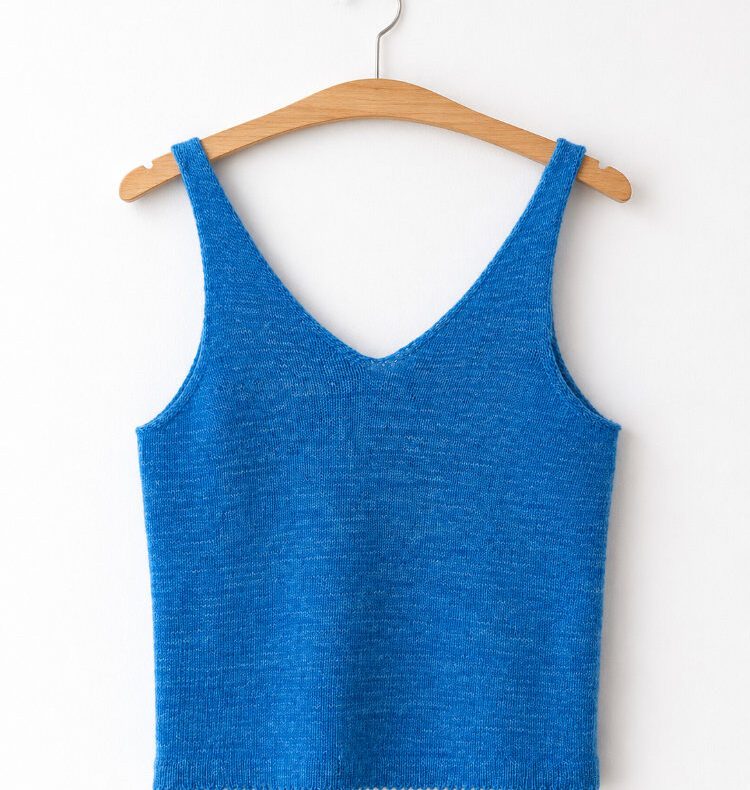 Blaues handgestricktes Niine Top mit V-Ausschnitt und breiten Trägern, nahtlos von oben gestrickt. Das schlichte Sommertop hängt an einem Holzbügel vor einer weißen Wand.