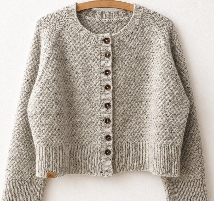 Greetl Cardigan aus meliertem graubeigem Garn auf einem Holzbügel an einer hellen Wand, mit Rundhalsausschnitt, Rippenblenden und Knopfleiste.