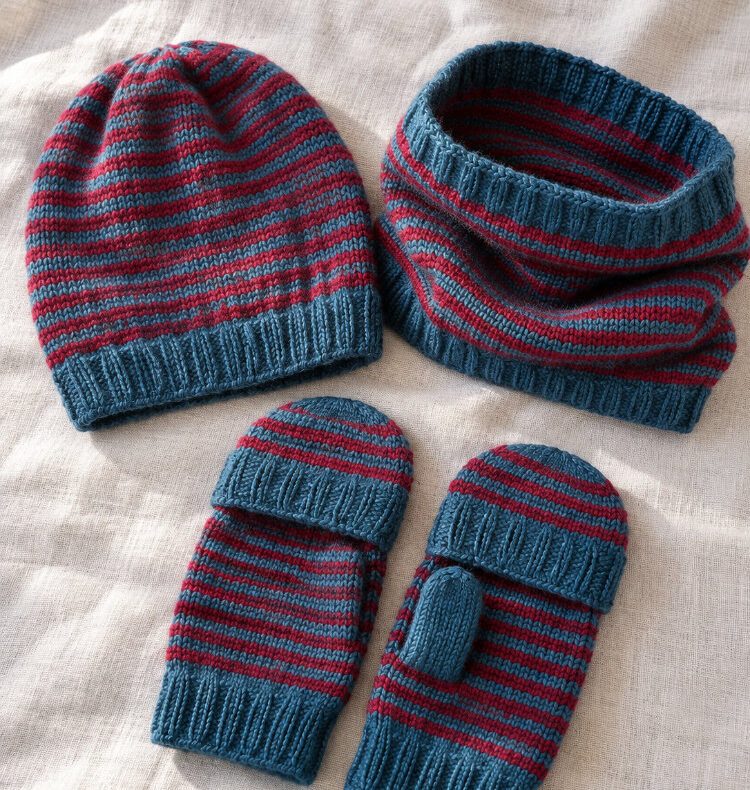 Gestricktes Truudi Set mit Mütze, Halswärmer und Handschuhen mit Fingerklappen in Blau und Rot auf hellem, neutralem Stoff.