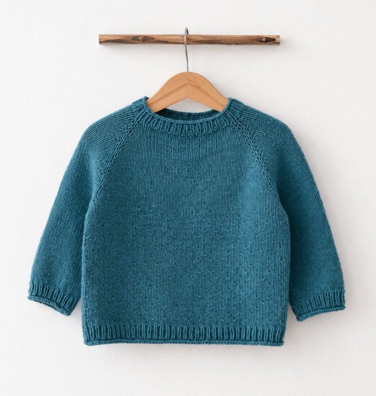 Türkisblauer Kindersweater auf Holzbügel vor heller Wand, mit Raglanzunahmen, kurzen Ärmeln und Rippenabschlüssen.