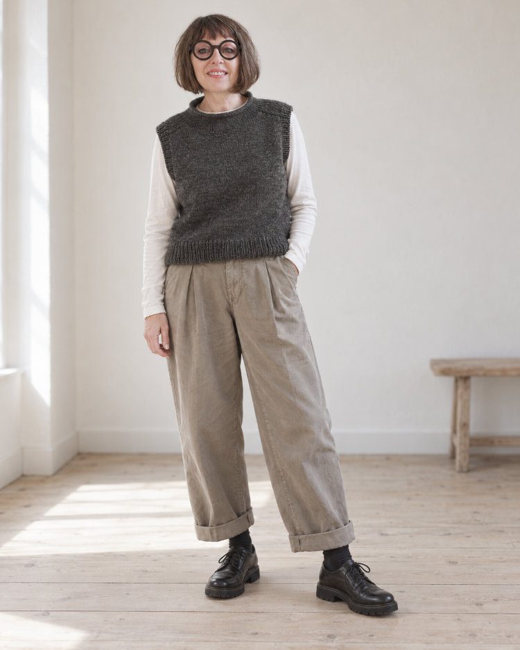 Frau trägt den anthrazitfarbenen Truudi Vestover über einem hellen Langarmshirt zu weiter Bundfaltenhose und schwarzen Schnürschuhen.