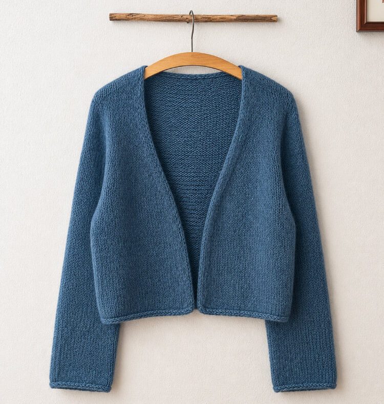 Uukii Cardigan in Petrolblau, handgestrickt mit offenem Vorderteil und langen Ärmeln. Der nahtlos von oben gestrickte Cardigan hängt auf einem Holzbügel vor einer schlichten weißen Wand und zeigt seine klare, minimalistische Form sowie das ruhige Maschenbild.