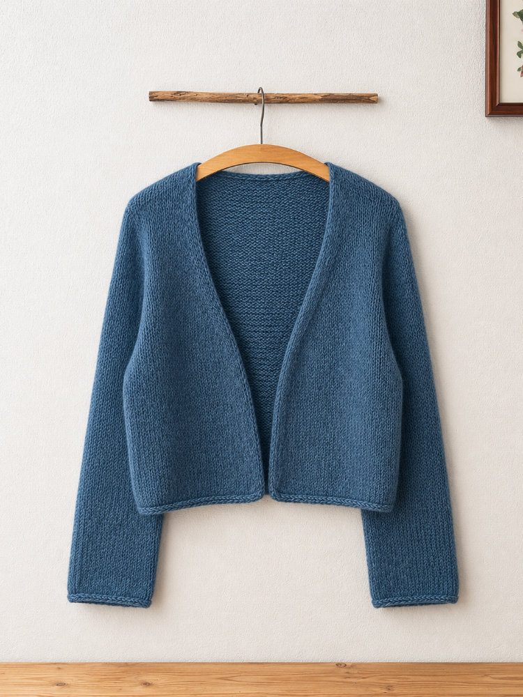 Uukii Cardigan in Petrolblau, handgestrickt mit offenem Vorderteil und langen Ärmeln. Der nahtlos von oben gestrickte Cardigan hängt auf einem Holzbügel vor einer schlichten weißen Wand und zeigt seine klare, minimalistische Form sowie das ruhige Maschenbild.