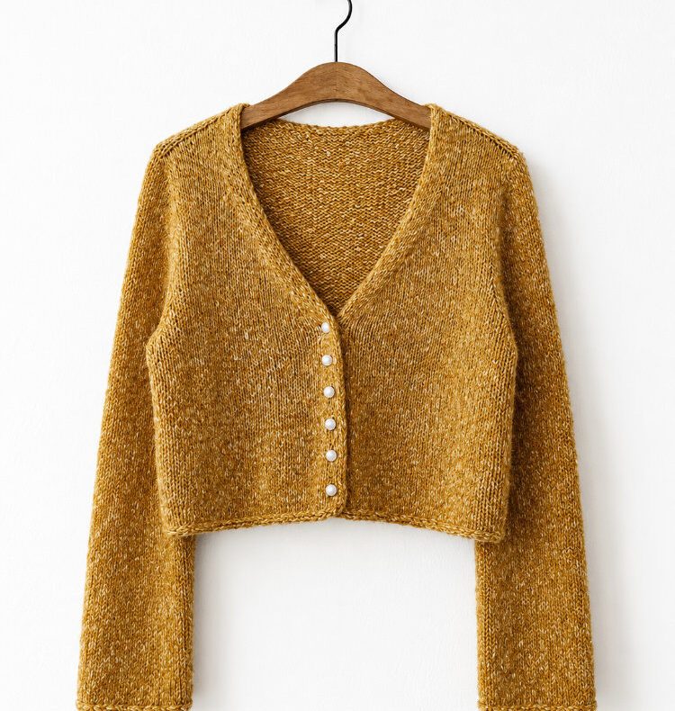 Handgestrickter Uukii Cardigan in warmem Senfgelb aus Tweedgarn mit V-Ausschnitt und Knopfleiste. Der nahtlos von oben gestrickte Cardigan hängt auf einem Holzbügel vor einer weißen Wand und zeigt die kurze, moderne Passform.