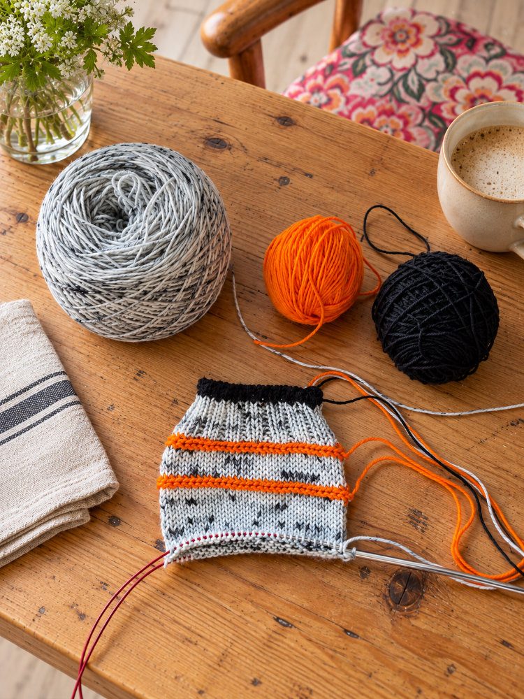 Socke in Grau, Schwarz und Orange mit passenden Garnknäueln und Rundstricknadel auf einem Holztisch.