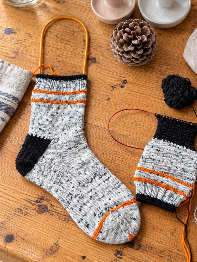 Gestrickte Pippa Socke und zweite Socke auf der Rundstricknadel mit Garnknäueln und Projektbeutel auf einem Holztisch.