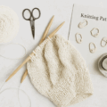 Strickprojekt mit hellem Garn auf rustikalem Holzuntergrund, daneben ein florales Notizbuch – stilles Bild zum Thema „Stricken auf Reisen“
