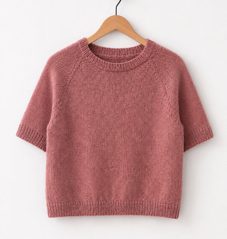 Altrosa handgestricktes Doorli Shortsleeve mit modifizierter Raglannaht auf einem Holzbügel vor weißer Wand.