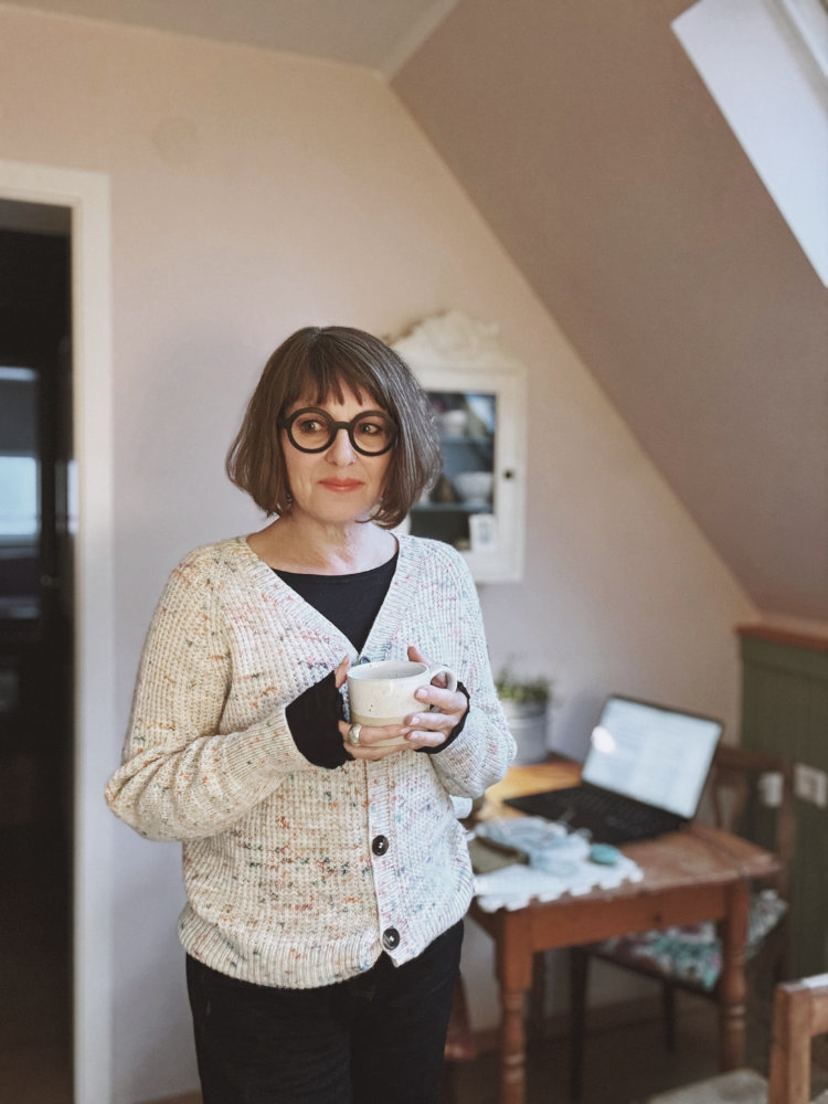 Tavalli Kollektion: Frau hält ein gestricktes Tavalli Projekt aus hellem Garn mit bunten Tweed-Sprenkel in den Händen, rosa p. Kollektion