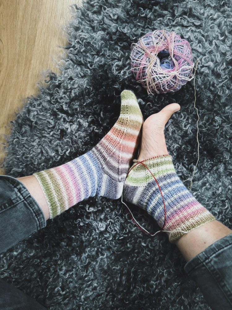 Gestrickte Tavalli Socks mit buntem Streifenmuster am Fuß, angezogen auf grauem Fellteppich, daneben ein Knäuel Wolle, Design von rosa p.