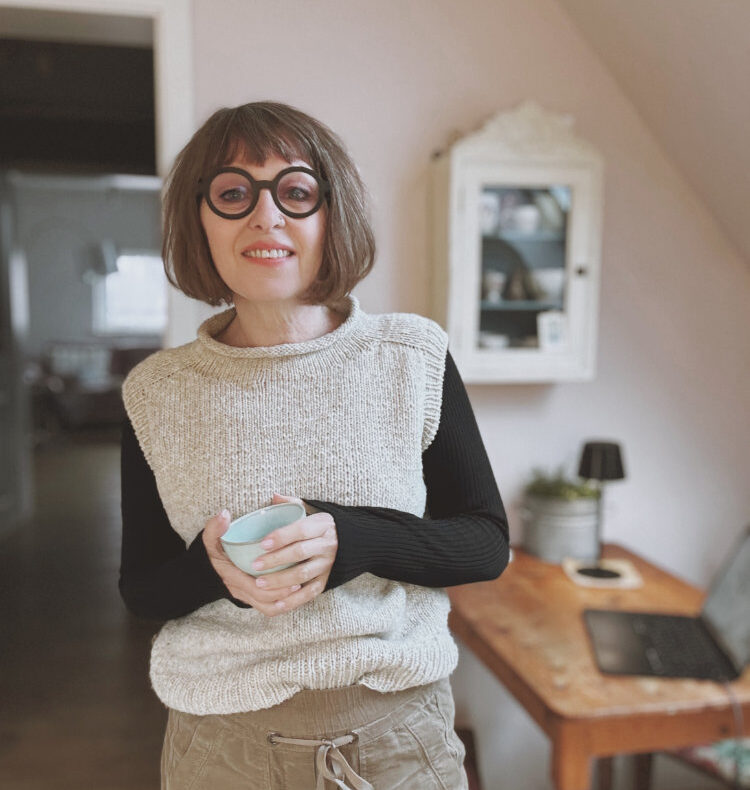 Truudi Sweater gestrickt aus De Rerum Natura Gilliatt in Dunkelblau – klassischer Pullover mit Rundhalsausschnitt, getragen von Frau mit Brille und Kaffeetasse in der Hand