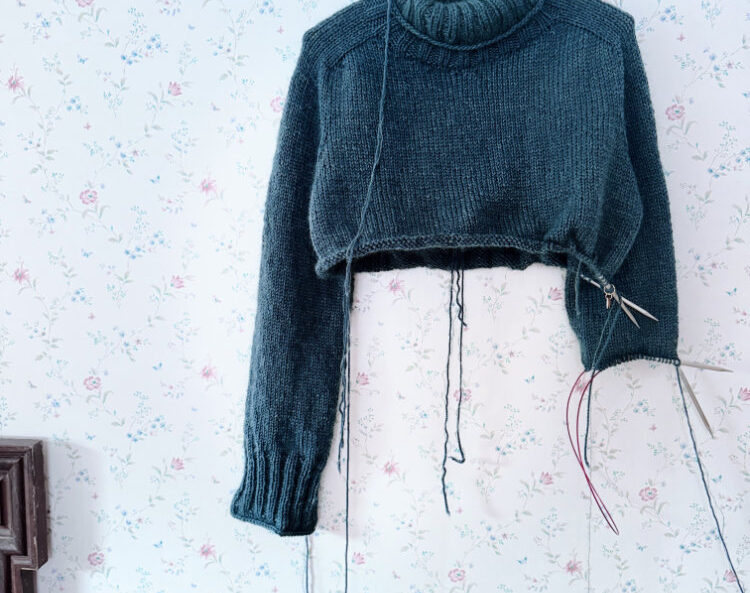 Truudi Sweater gestrickt aus Drops Daisy und Kid-Silk – klassischer Pullover mit Rundhalsausschnitt, glatt rechts gestrickt, in Dunkelpetrol auf Kleiderbügel vor gemusterter Tapete.