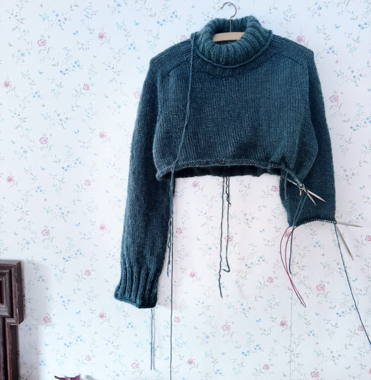 Truudi Sweater gestrickt aus Drops Daisy und Kid-Silk – klassischer Pullover mit Rundhalsausschnitt, glatt rechts gestrickt, in Dunkelpetrol auf Kleiderbügel vor gemusterter Tapete.