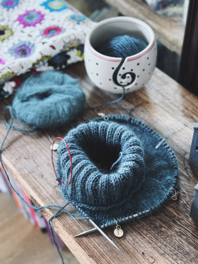 Truudi Kollektion – Strickprojekt in Arbeit mit blaugrauer Wolle auf dem Tisch. Nahtlos gestrickt, klassisches rosa p.-Design mit klarer Linie.