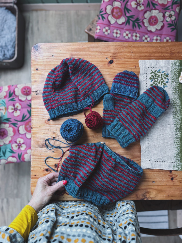 Truudi Set bestehend aus Truudi Sweater und Truudi Vestover – gestrickt in warmen Beerentönen, liegend auf Holztisch mit Strickzubehör und Garnknäueln.
