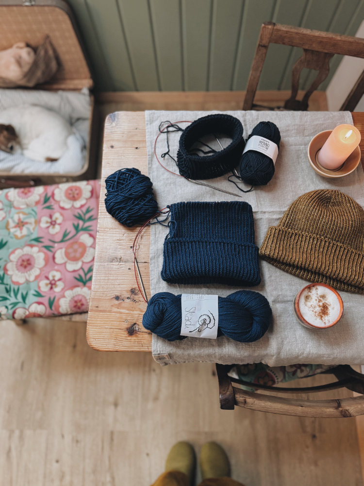 Strickdetails auf einem Holztisch: Garnknäuel, ein kleines zweifarbiges Set aus Mütze und Hose