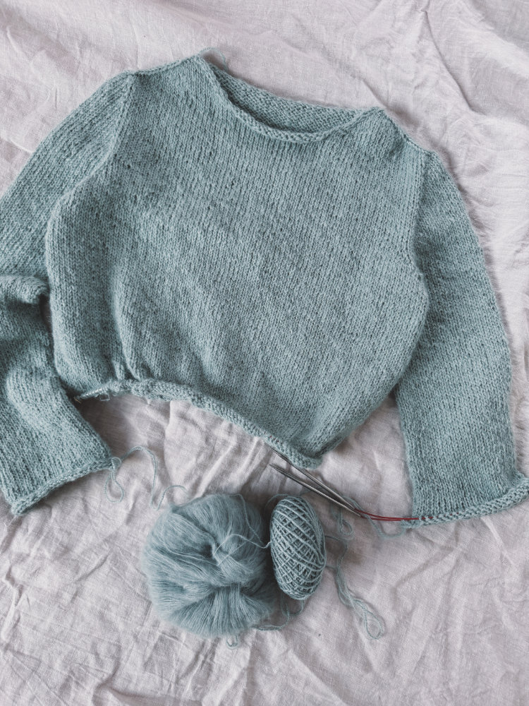 Uukii Sweater in hellem Grün, flach liegend auf einem Holztisch, mit sichtbarer Rundpasse und weichem Maschenbild.