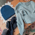 Hellgrauer Uukii Sweater und dunkelblauer Zoora Hat liegen gefaltet auf einem Tisch – winterliche Strickprojekte für die Zeit zwischen den Jahren.