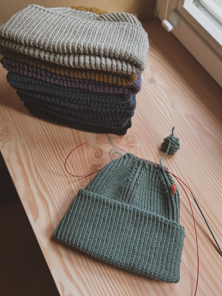 Zoora Hats auf einem Holztisch mit Stricknadeln – Beispiel für Mützensitz mit negativer Bequemlichkeitszugabe.