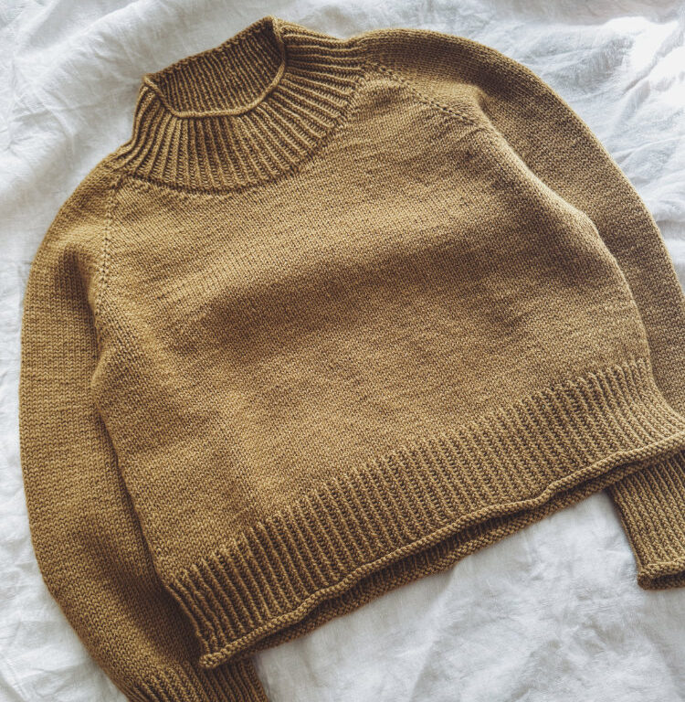 : Zoora Sweater – sandfarbener, handgestrickter Raglanpullover mit Turtle-Neck, flach liegend auf hellem Stoff.