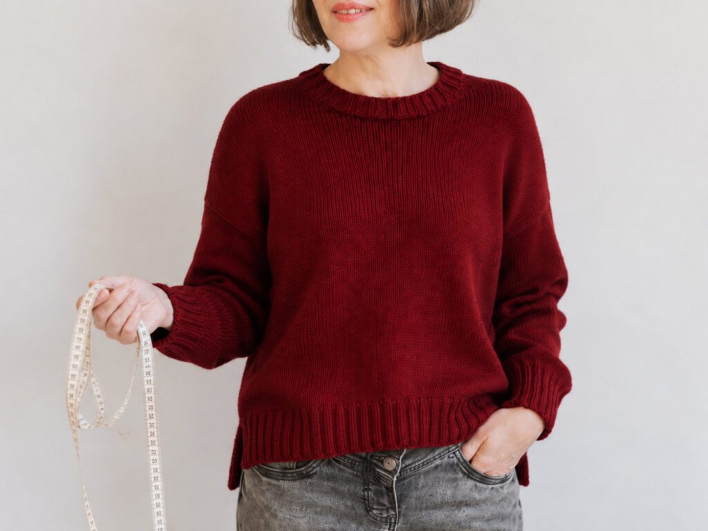 Kaiili Jumper in Dunkelrot, getragen mit Maßband zur Größenwahl einer Strickanleitung.