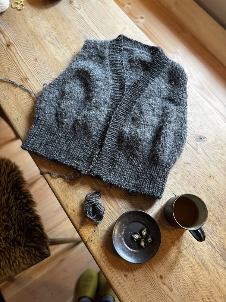 Erikka Vest in Dunkelgrau – Stillleben mit Kaffee und Garn auf Holztisch.