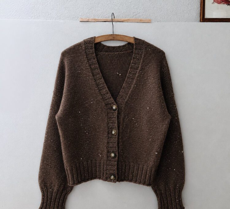 Brauner Cardigan aus Tweed mit V-Ausschnitt und Knopfleiste an einem Holzbügel vor einer hellen Wand.