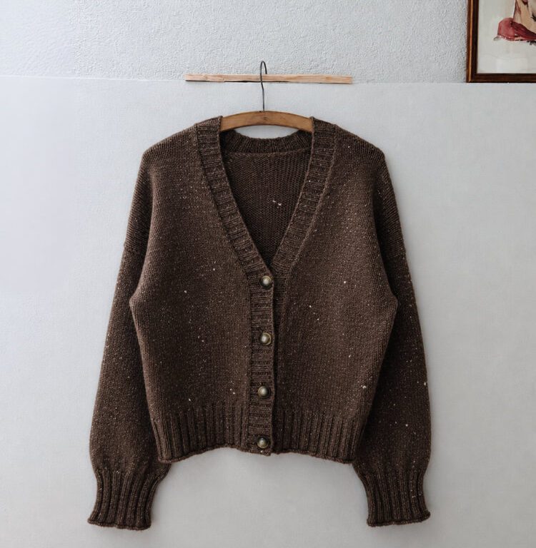Brauner Cardigan aus Tweed mit V-Ausschnitt und Knopfleiste an einem Holzbügel vor einer hellen Wand.