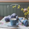 Frühling bei rosa p. – Kaffeetasse auf Tisch, daneben Garnknäuel und Stricknadeln (Stillleben).
