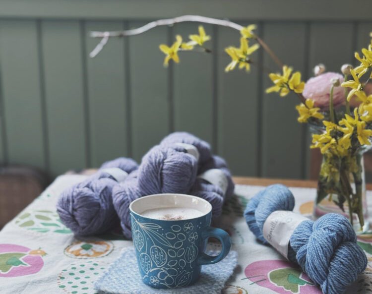 Frühling bei rosa p. – Kaffeetasse auf Tisch, daneben Garnknäuel und Stricknadeln (Stillleben).