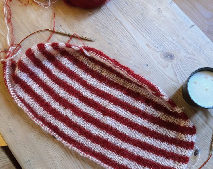 Roosi Blouse in Arbeit – rot-weiß gestreiftes Strickstück auf Holztisch, mit Maschenmarkierern.