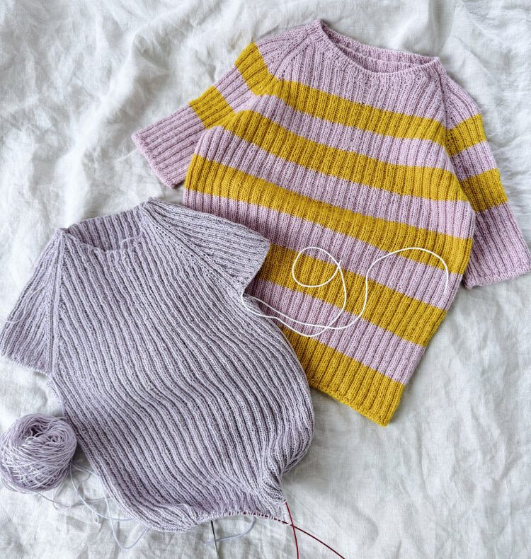 Riika Shirt – zwei handgestrickte Shirts in kraus rechts: ein helles Shirt und ein gelb-weiß gestreiftes Shirt, als Flatlay auf hellem Stoff.