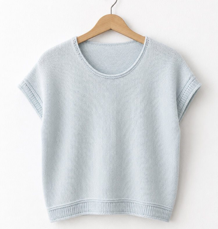 Hellblaues Heedi Tee aus glattem Strick an einem Holzkleiderbügel vor einer weißen Wand. Nahtlos von oben gestricktes Sommershirt mit europäischer Schulter, schmalem Rundhalsausschnitt, kurzen Ärmeln und schlichtem Rollsaum.