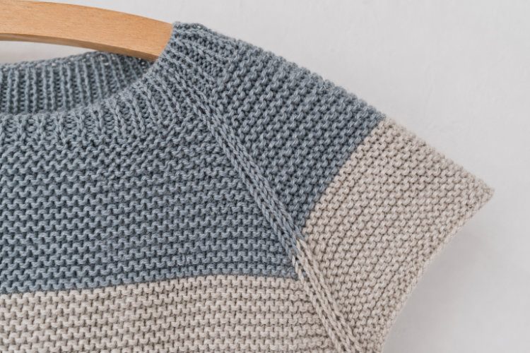 Detailaufnahme einer deutlich sichtbaren Raglanlinie an einem blau-grau und cremefarben gestrickten Modell. Die diagonale Linie zwischen Körper und Ärmel zeigt den typischen Aufbau der Raglankonstruktion.