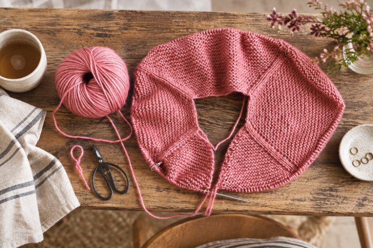 Rosafarbenes Strickstück mit sichtbaren Raglanlinien, passendem Wollknäuel und kleiner Schere auf einem Holztisch.